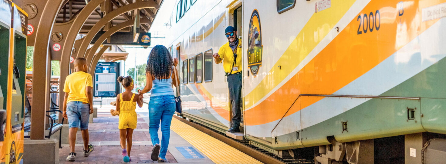 Woman with 2 children walking on platform at Longwood Station with SunRail Engineer waving from train.