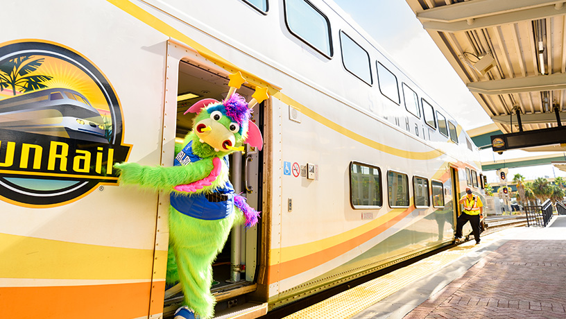 Landing Page image - Orlando Magic's Stuff the Magic Dragon standing in the doorway of a parked SunRail train, as it awaits passengers to board at the Church Street Station.