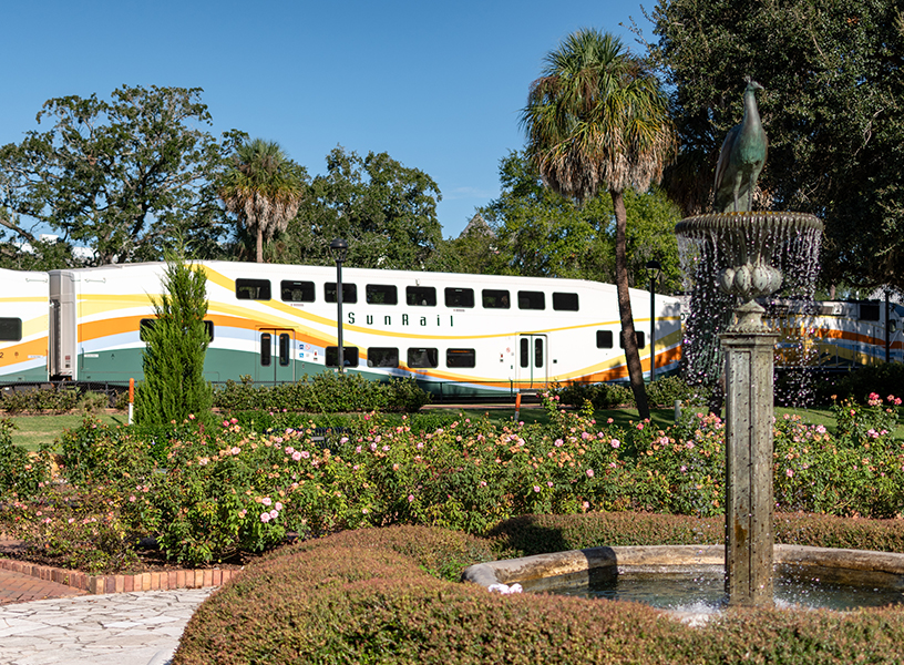 The Winter Park Peacock Fountain sits in the center of Park Avenue's rose garden with a SunRail train in the backgroun.