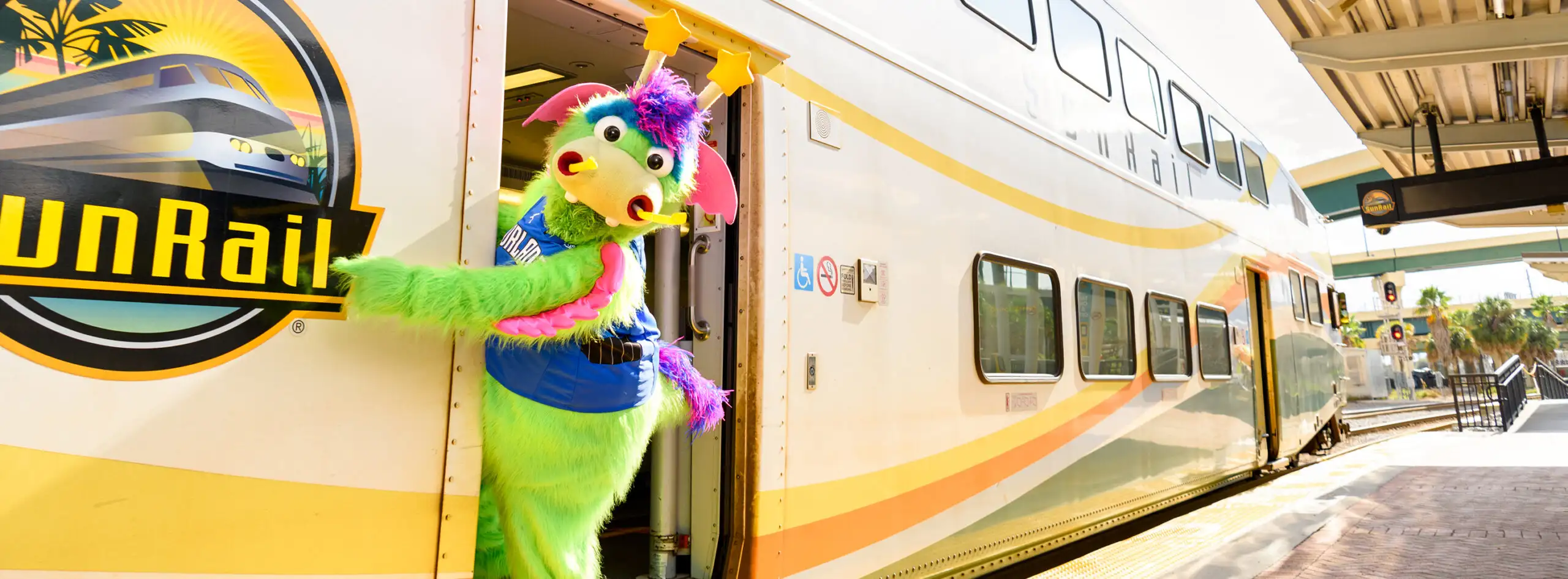 Masthead image - Orlando Magic's Stuff the Magic Dragon standing in the doorway of a parked SunRail train, as it awaits passengers to board at the Church Street Station.